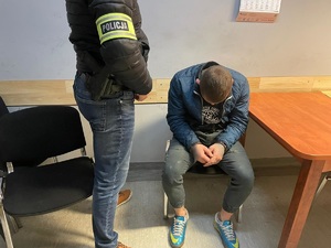zdjęcie przedstawiające policjanta, stojącego przy siedzącym na krześle zatrzymanym