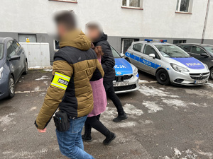 zdjęcie przedstawia policjantów z zatrzymaną kobietą idących przez parking komendy gdzie stoją policyjne radiowozy