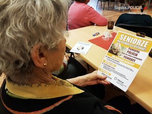 zdjęcie poglądowe - starsza pani siedzi przy stoliku czytając ulotkę dotyczącą przestępstw na seniorach