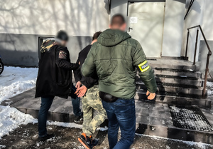 zdjęcie przedstawia policjantów prowadzących zatrzymanego z parkingu do budynku komendy
