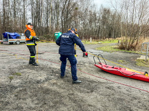 zdjęcie przedstawia policjant i strażak ciągnący linę do której przyczepiony jest pozorant na stawie