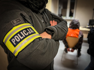 zdjęcie przedstawia policjantów po cywilu z opaską policja na ręce, w tle widać siedzącego zatrzymanego