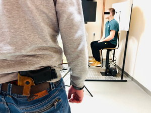 zdjęcie w pomieszczeniu technika kryminalistyki w trakcie wykonywania zdjęć do policyjnej bazy zatrzymanemu sprawcy - policjant po cywilu stoi, a za nim na krześle maszyny fotograficznej zatrzymany