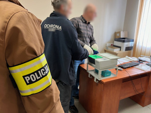 zdjęcie przedstawia policjanta kryminalnego w opasce z napisem policja oraz zatrzymanego i technika kryminalistyki pobierającego odciski palców