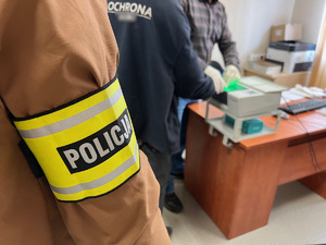 zdjęcie przedstawia policjanta kryminalnego w opasce z napisem policja oraz zatrzymanego i technika kryminalistyki pobierającego odciski palców