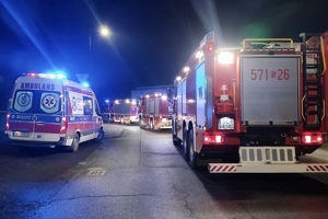 zdjęcie przedstawiające ulicę dojazdową do miejsca zdarzenia i stojące po obu stronach pojazdy służb ratunkowych z włączonymi niebieskimi światłami błyskowymi
