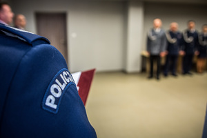 sala narad, widoczna ręka prawa policjanta odczytującego rozkazy personalne, w tle widoczne sylwetki stojących policjantów