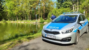 Na zdjęciu znajduje się radiowóz na drodze szutrowej, 1 policjant, w tle zbiornik wodny i las