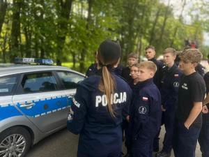 Na zdjęciu znajduje się jedna policjantka z uczniami liceum w trakcie rozmowy na tle radiowozu policyjnego