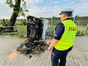 zdjęcie z miejsca zdarzenia - leżący na boku samochód dostawczy, obok drzewo w które uderzył, stojący przy nim policjant