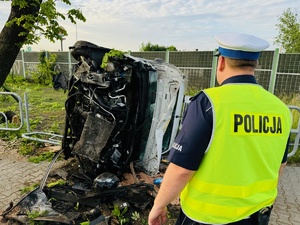 zdjęcie z miejsca zdarzenia - leżący na boku samochód dostawczy, obok drzewo w które uderzył, stojący przy nim policjant