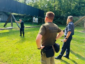 zdjęcie przedstawia zajęcia na strzelnicy otwartej podczas których instruktor wyjaśnia przebieg ćwiczenia, policjanci stoją słuchając