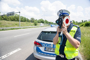 zdjęcie przedstawia policjanta z drogówki z miernikiem prędkości stojącego przy trzypasmowej drodze