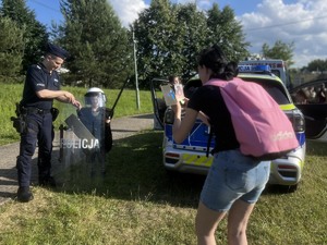 Na zdjęciu dzielnicowy pomaga przytrzymać chłopcu tarczę z napisem policja. Chłopiec stoi na tle radiowozu a kobieta robi mu zdjęcie