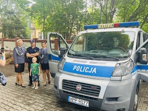 Na zdjęciu policjantki w umundurowaniu galowym, policjant w umundurowaniu służbowym i dziecko, pozują do zdjęcia przy radiowozie