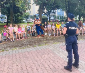 Na zdjęciu maskotka Sierżant Ruda siedzi obok dzieci, policjantka prowadzi pogadankę