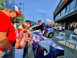 zdjęcie z festynu przedstawiające przygotowane przez policjantów stoisko profilaktyczne, policyjnego kadrowca i oglądające dzieci z rodzicem