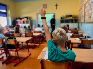 zdjęcie z wnętrza klasy, widoczne są siedzące w ławkach dzieci, a jedno z nich podnosi rękę aby udzielić odpowiedzi