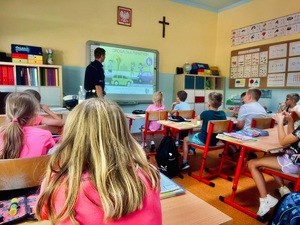 zdjęcie z wnętrza klasy, widoczne są siedzące w ławkach dzieci i policjant opowiadający o tym co widać na wyświetlanej prezentacji