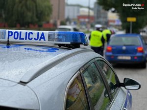 zdjęcie przedstawiające radiowóz policyjny stojący przy głównej ulicy miasta i dwóch policjantów z drogówki kontrolujących samochód osobowy stojący przed radiowozem