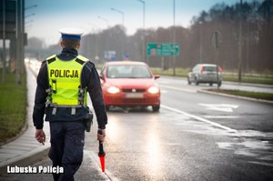 zdjęcie poglądowe z archiwum policji - policjant z drogówki stojący przy mokrej drodze, gdzie jedzie czerwony samochód z włączonymi światłami