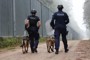 zdjęcie przedstawia dwóch policjantów z psami służbowymi idących wzdłuż zapory na granicy Polcki z Białorusią