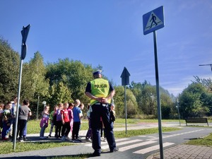 Dzieci wraz z umundurowanym policjantem w miasteczku ruchu drogowego, ćwiczą przejście przez jezdnię