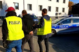 zdjęcie poglądowe przedstawia policjantów prowadzących zatrzymanego przez policyjny parking, widoczne są stojące pojazdy policji i budynek w tle - zdjęcie z archiwum rudzkiej policji