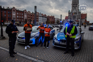 zdjęcie z akcji Świeć przykładem noś odblaski przedstawiające policjantów i wolontariuszy z odblaskami na tle dwóch radiowozów stojących na głównym placu miasta