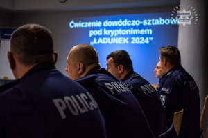 zdjęcie przedstawia salę odpraw komendy, siedzących policjantów, a na ekranie wyświetlane założenie ćwiczeń dowódczo-sztabowych Ładunek 2024