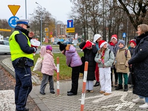 zdjęcie przedstawia policjanta i grupkę uczniów w czapkach mikołaja przy przejściu dla pieszych