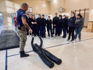 Na zdjęciu grupa uczniów i instruktor, który prezentuje ćwiczenie z manekinem zapaśniczym