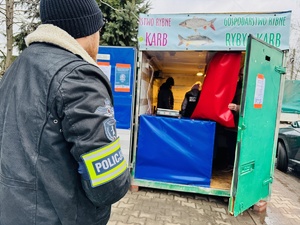 zdjęcie przedstawia kontrolę punktu sprzedaży karpia, policjant stojący przed punktem i inspektorzy weterynarii rozmawiający z sprzedawcą