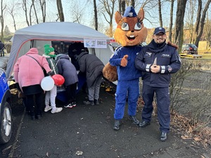 zdjęcie przedstawia policjanta z maskotką policji przy stoisku z osobami zainteresowanymi ulotkami