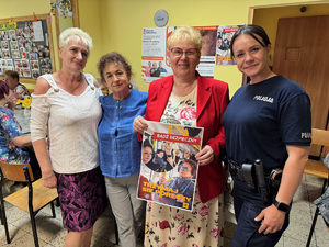zdjęcie przedstawia pomieszczenia koła seniorów, gdzie policjantka stoi z trzema kobietami, jedna z nich trzyma plakat akcji