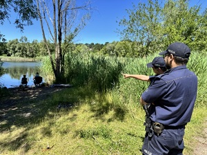 zdjęcie przedstawia policjantów w trakcie kontroli stawu, policjantka wskazuje na chłopca z mężczyzną łowiących ryby