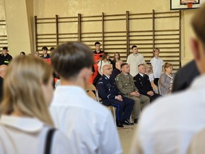 zdjęcie przedstawia komendantów służb mundurowych siedzących na krzesłach w sali gdzie odbywa się rozpoczęcie roku szkolnego, na zdjęciu widać stojących uczniów pierwszej klasy