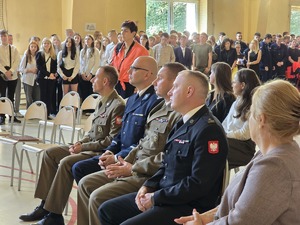 zdjęcie przedstawia komendantów służb mundurowych siedzących na krzesłach w sali gdzie odbywa się rozpoczęcie roku szkolnego, w tle widać stojących uczniów