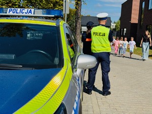 zdjęcie przedstawia policjanta i policjantkę obok szkoły gdzie idzie wychowawca z dziećmi