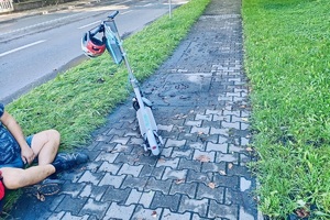 Na zdjęciu hulajnoga z zawieszonym kaskiem na kierownicy, obok siedzi mężczyzna