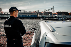 zdjęcie przedstawia policjanta przy radiowozie obserwującego teren bocznicy kolejowej z wagonami towarowymi