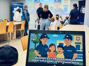 zdjęcie przedstawia laptopa z wyświetloną grafiką pokazującą pracę policjantów, a w tle widać jak dzieci rozmawiają z policjantami