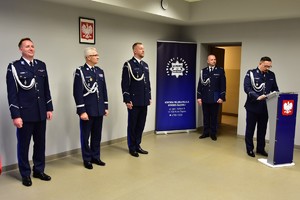zdjęcie przedstawia salę w której odbywa się uroczysta zbiórka, na fotografii widać Komendanta Wojewódzkiego Policji, dwóch zmieniających się komendantów i policjantów pełniących funkcję lektorów