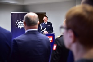 zdjęcie przedstawia salę w której odbywa się uroczysta zbiórka, na fotografii widać zebranych gości i między nimi przemawiającego zdającego stanowisko komendanta