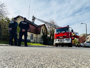 zdjęcie przedstawia dwóch policjantów stojących na ulicy, w tle strażacka drabina i budynek komisariatu