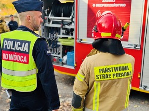 zdjęcie przedstawia kierującego akcją ze strony policji rozmawiającego ze strażakiem na tle wozu straży pożarnej