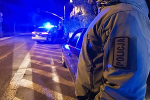 zdjęcie przedstawia policjanta kryminalnego przy samochodzie, a w tle widać radiowóz oznakowany z włączonymi światłami błyskowymi