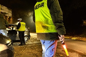 zdjęcie przedstawia policjantów kontrolujących samochód i obok policjanta kryminalnego w kamizelce odblaskowej z latarką w ręku