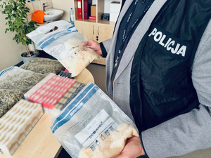 zdjęcie przestawia policjanta służby kryminalnej trzymającego w rękach worki z narkotykami, na stole obok leżą kolejne worki z narkotykami i paczki papierosów