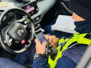 zdjęcie przedstawia policjanta z drogówki siedzącego w radiowozie podczas kontroli danych na urządzeniu mobilnym